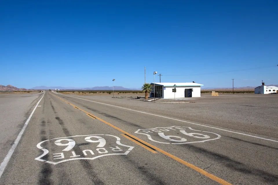 Low shot of Route 66 signs on asphalt with small abandoned building on roadside on old Route 66 near Amboy, California.